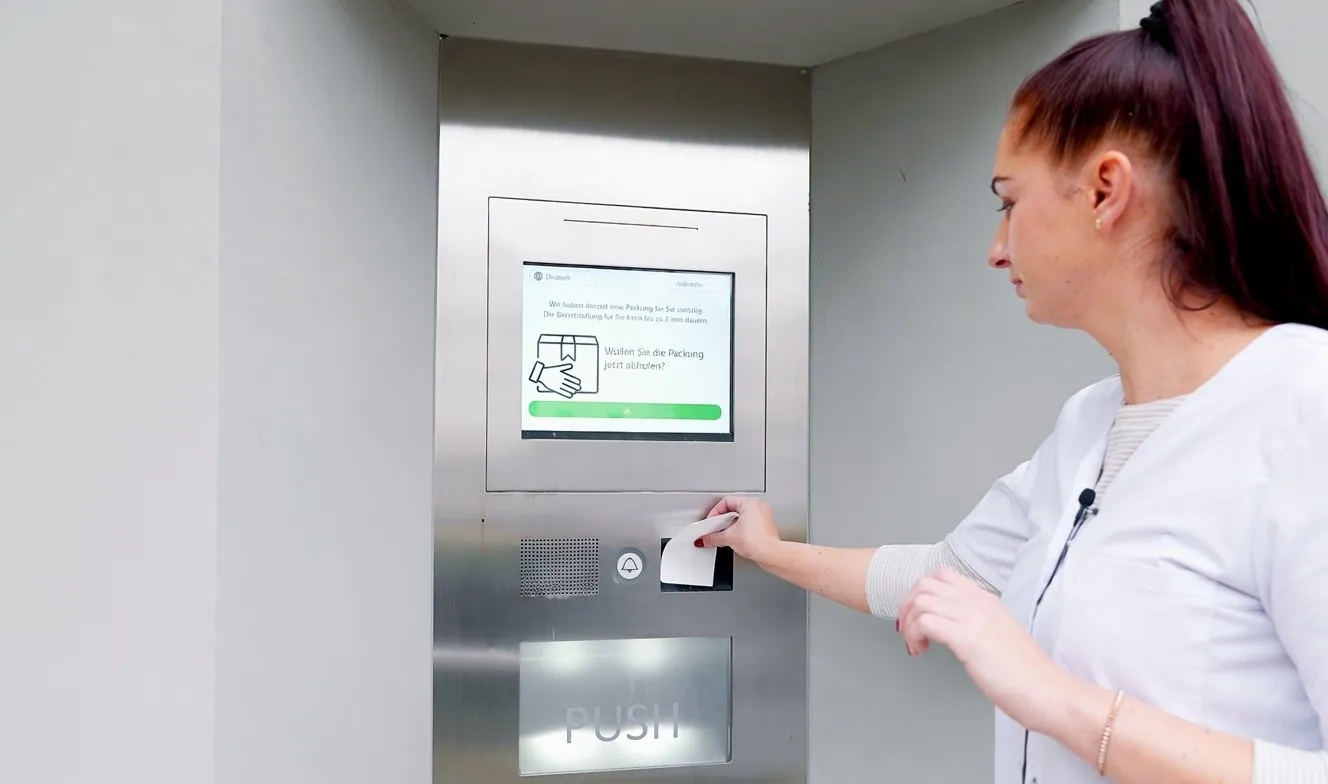 Mitarbeiterin bedient den Bildschirm des Abholautomaten und hält den Abholcode in der Hand
