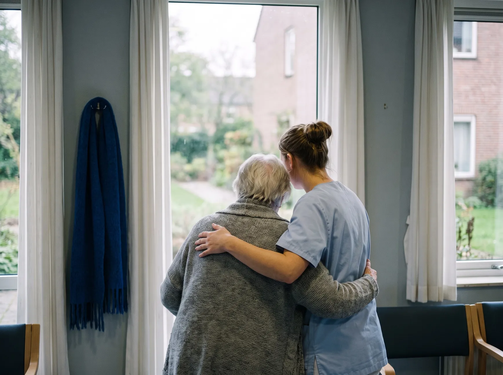 Pflegerin steht mit dem Arm um eine Bewohnerin am Fenster eines Pflegeheims